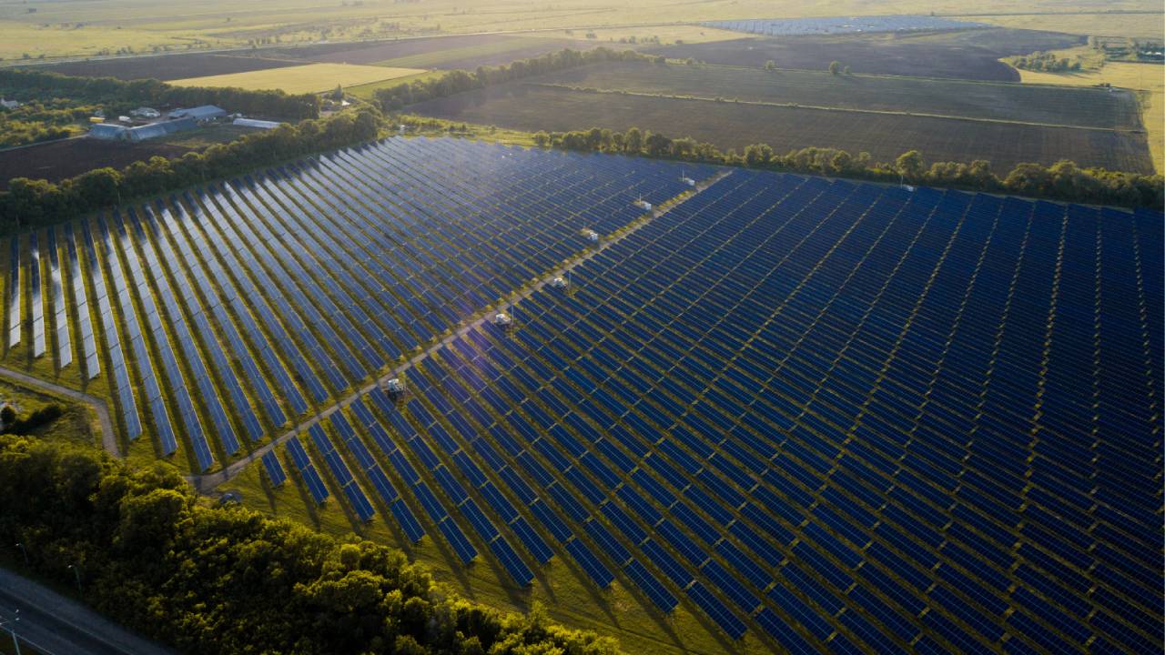 Vista aérea de uma usina de energia solar com placas fotovoltaicas e áreas de vegetação ao pôr do sol. 